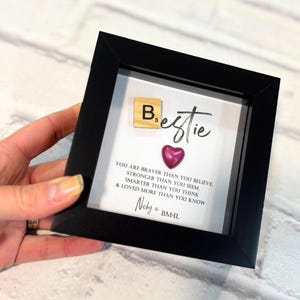 May include: A black framed picture with a white background and the text "Bestie" in black script. The text "You are braver than you believe, stronger than you seem, smarter than you think, & loved more than you know" is printed below. A small wooden tile with the letter "B" and the number "3" is placed above the word "Bestie". A small pink heart is placed below the word "Bestie". The text "Neby BM4L" is printed below the quote.