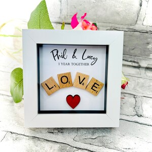 May include: White frame with a black mount and a white background. The text "Phil & Lucy 1 Year Together" is written in black cursive. The word "LOVE" is spelled out with wooden letter tiles. A red heart is in the centre of the tiles.