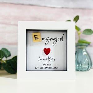 Könnte beinhalten: Ein weißer Rahmen mit schwarzem Passepartout und weißem Hintergrund. Der Text "E, ngaged" ist in schwarzer Schrift geschrieben. Ein rotes Herz und eine Holzkachel mit dem Buchstaben "E" sind auf dem Hintergrund platziert. Der Text "Lee and Kelly" ist unter dem Herzen geschrieben. Der Text "Dubai" ist unter den Namen geschrieben. Das Datum "13th September 2024" ist unter der Stadt geschrieben.