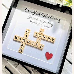 Op de afbeelding: Een ingelijst huwelijkscadeau met de woorden "Gefeliciteerd Sarah & Jonny" en "Getrouwd 13 september". Houten letters spellen "GETROUWD" en "LIEFDE", met een rood hart.