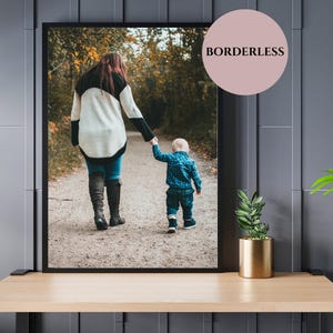May include: Framed photograph of a person and a child walking on a path. The person wears a black and white sweater, blue jeans, and black boots. The child wears a blue plaid shirt and jeans. The image is displayed on a wooden table.