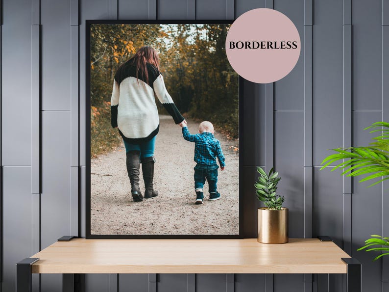 May include: Framed photograph of a person and a child walking on a path. The person wears a black and white sweater, blue jeans, and black boots. The child wears a blue plaid shirt and jeans. The image is displayed on a wooden table.