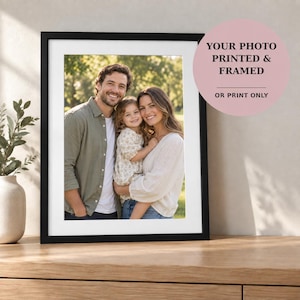May include: A black rectangular picture frame containing a photograph of a man, a woman, and a young child standing outdoors. The frame includes a white mat border. A pink circular graphic overlay displays the text: YOUR PHOTO PRINTED & FRAMED OR PRINT ONLY.