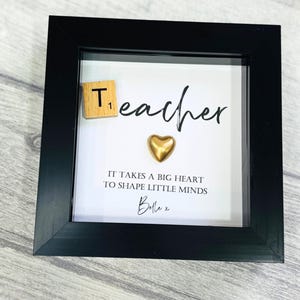 Puede incluir: Una imagen enmarcada en negro con fondo blanco. La imagen dice "Teacher" con un corazón dorado y un azulejo de madera con el número "1" en él. El texto debajo del corazón dice "Se necesita un gran corazón para moldear las mentes pequeñas" y está firmado "Belle x".