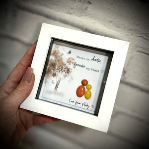 Puede incluir: Imagen enmarcada en blanco con una pintura de acuarela de un árbol y las palabras "Always my Auntie, forever my friend" con un corazón. Tres piedras de vidrio de colores están dispuestas para parecer una familia. El texto "Love from Nicky" está escrito debajo de las piedras.