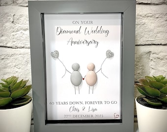 May include: A framed art piece celebrating a Diamond Wedding Anniversary. The artwork features two figures made of gray and pink stones, holding hands and balloons. The text reads "On Your Diamond Wedding Anniversary", "60 Years Down, Forever To Go", "Chris & Lisa", and "22nd December 2023".
