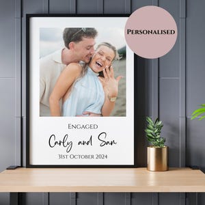 May include: A framed photo print featuring a couple celebrating an engagement. The photo is set against a white background with the words "ENGAGED" and the names "Carly and Sam" along with the date "31st October 2024". A pink circle with the word "PERSONALISED" is in the upper right corner.