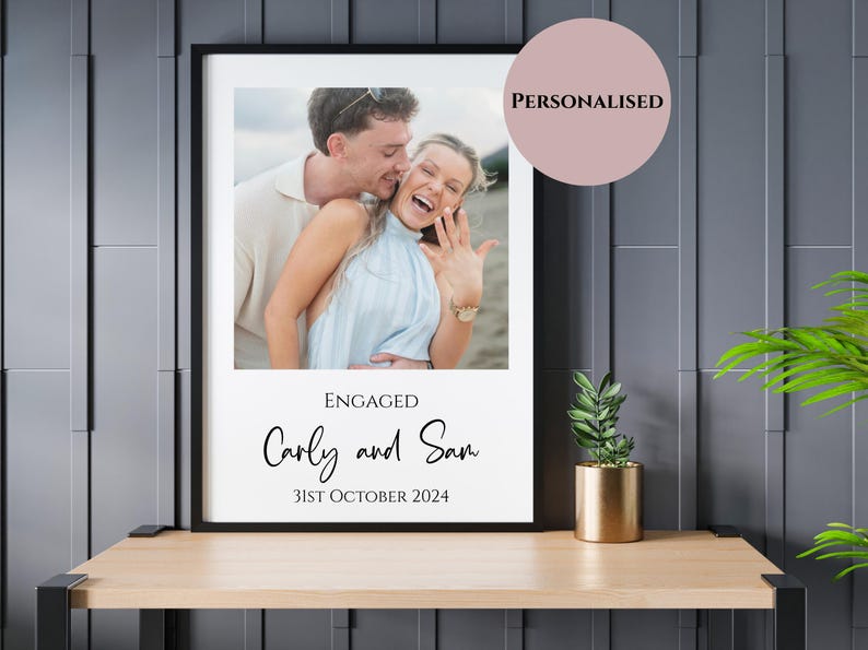 May include: A framed photo print featuring a couple celebrating an engagement. The photo is set against a white background with the words "ENGAGED" and the names "Carly and Sam" along with the date "31st October 2024". A pink circle with the word "PERSONALISED" is in the upper right corner.