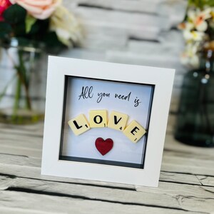 Può includere: Immagine incorniciata bianca con il testo "All you need is" e la parola "LOVE" scritta con tessere da Scrabble in una forma curva. Un cuore rosso si trova sotto le tessere.