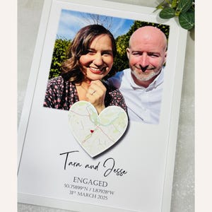 Könnte beinhalten: Ein weiß gerahmter Foto-Druck mit einem lächelnden Paar und einer herzförmigen Karte. Der Text lautet "Tara and Jesse", "ENGAGED", das Datum und die Koordinaten. Der Rahmen ist weiß.