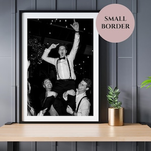 May include: Black and white framed photograph of three people celebrating. One person is being held up on the shoulders of two others, with arms raised in the air. The image is displayed on a wooden table with a gold pot and plant.