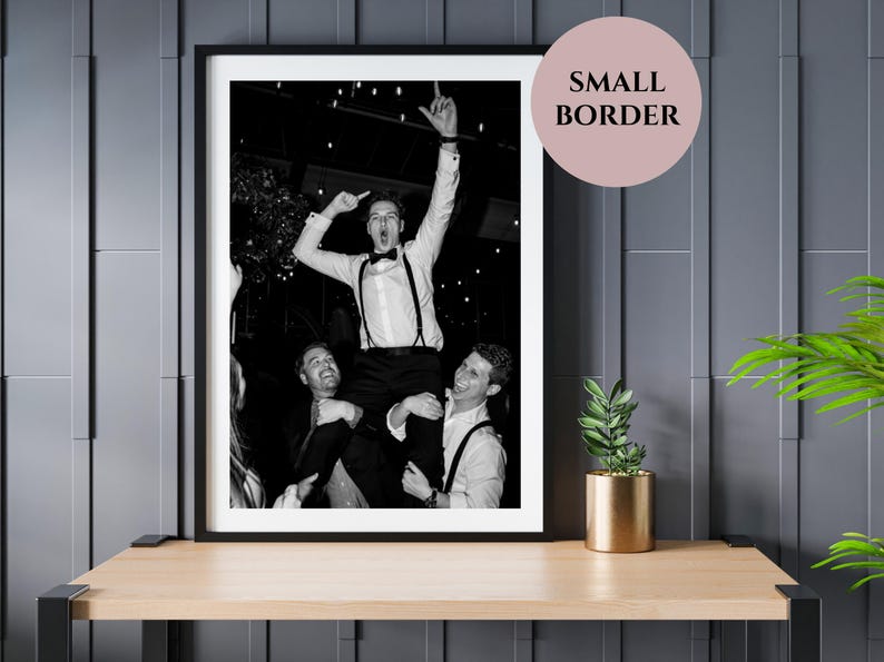 May include: Black and white framed photograph of three people celebrating. One person is being held up on the shoulders of two others, with arms raised in the air. The image is displayed on a wooden table with a gold pot and plant.