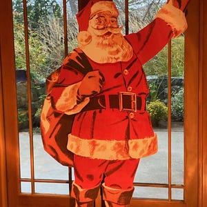 May include: A large, flat, die-cut cardboard cutout of Santa Claus wearing a traditional red suit with white fur trim, a black belt with a gold buckle, and black boots. Santa is depicted with a white beard, carrying a large sack over one shoulder.