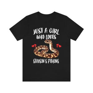 May include: Black t-shirt featuring the text "JUST A GIRL WHO LOVES STIMSON'S PYTHONS" in white. A brown and tan snake graphic is centered below the text, with red heart graphics on either side.