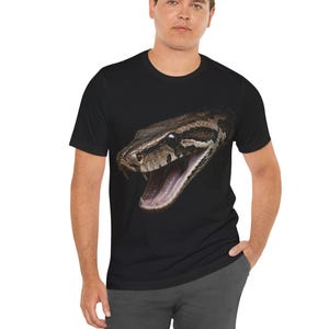 以下が含まれることがあります： 黒の半袖Tシャツで、口を大きく開けた茶色と黒のヘビの頭のリアルなグラフィックプリントが大きく施されています。口の中には牙が見え、衣服の前面中央に配置されています。