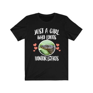 Puede incluir: Camiseta negra con texto blanco que dice "Just a girl who loves monitor lizards" con un gráfico circular de un lagarto monitor y corazones rosas.