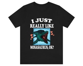 May include: Black t-shirt with a white graphic print of a green mosasaurus with an open mouth. The text "I just really like mosasaurus, ok?" is printed in white below the image.