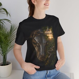May include: A black short-sleeve t-shirt featuring a large graphic print on the front. The design depicts a detailed close-up of a snake head emerging from a dense green forest landscape with a sunset visible in the background sky.