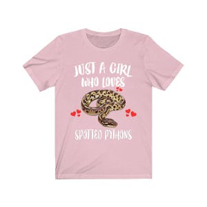 May include: A light pink t-shirt with the text "JUST A GIRL WHO LOVES SPOTTED PYTHONS" in white. The shirt features a graphic of a spotted python and red heart accents.