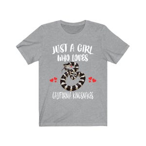 Op de afbeelding: Een heather grijze t-shirt met de tekst "JUST A GIRL WHO LOVES CALIFORNIA KINGSNAKES". De shirt heeft een afbeelding van een zwart, wit en bruin California Kingsnake met rode hart accenten. Het shirt is een casual kledingstuk.
