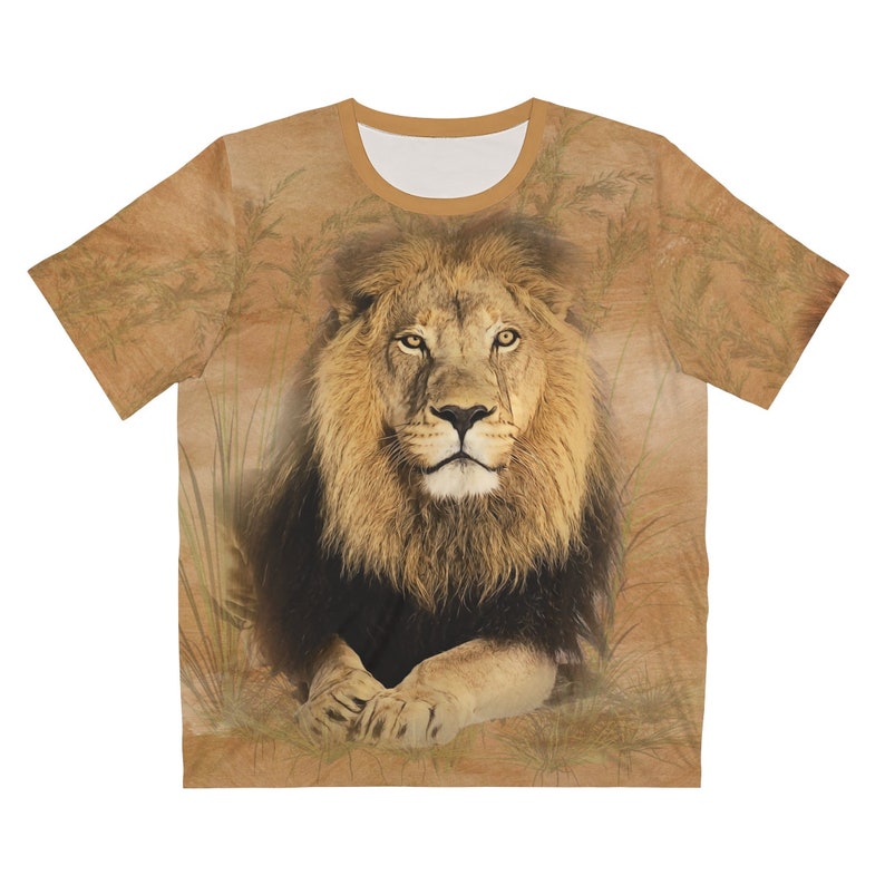 May include: A brown and tan t-shirt with a photorealistic image of a lion's face. The lion is lying down with its paws crossed and looking directly at the viewer. The background is a blurry image of tall grass.
