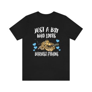 Puede incluir: Camiseta negra con el texto blanco "JUST A BOY WHO LOVES BURMESE PYTHONS". El diseño incluye un gráfico de una pitón birmana y corazones azules.