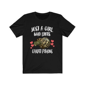 Op de afbeelding: Zwart T-shirt met witte tekst die "Just a girl who loves carpet pythons" luidt. Een cartoonafbeelding van een tapijtpython bevindt zich in het midden van het shirt. Roze harten bevinden zich rond de python.