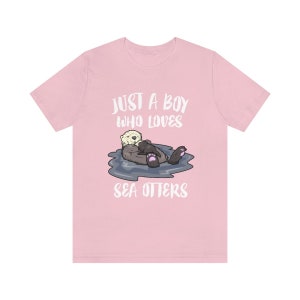 May include: Pink t-shirt with a white graphic of a sea otter holding a baby sea otter. The text "Just a boy who loves sea otters" is printed on the shirt.