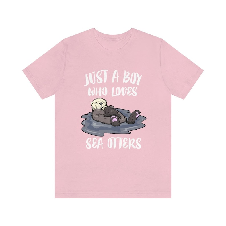 May include: Pink t-shirt with a white graphic of a sea otter holding a baby sea otter. The text "Just a boy who loves sea otters" is printed on the shirt.