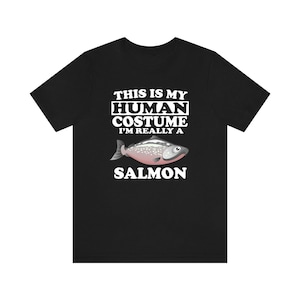 Op de afbeelding: Zwart t-shirt met de witte tekst "THIS IS MY HUMAN COSTUME I'M REALLY A SALMON". Een cartoon illustratie van een zalm staat onder de tekst, met roze, zilver en grijze kleuren.