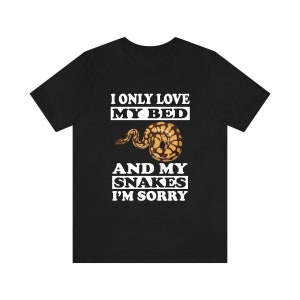 Op de afbeelding: Zwart T-shirt met de witte tekst "I ONLY LOVE MY BED AND MY SNAKES I'M SORRY". Een afbeelding van een bruine en gele slang is gecentreerd onder de tekst "MY BED".