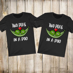 Peut inclure: Deux t-shirts noirs avec l'inscription "Two Peas in a Pod" au-dessus d'un dessin de deux petits pois dans une gousse avec un cœur rouge. Les t-shirts sont présentés sur un fond en bois.