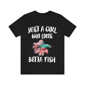 May include: A black short-sleeved t-shirt featuring white text that reads JUST A GIRL WHO LOVES BETTA FISH. Centered between the lines of text is a colorful illustration of a betta fish with a teal head and flowing pink and red fins.
