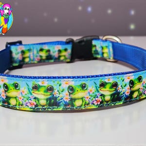 May include: A blue dog collar with a repeating pattern of smiling green frogs and flowers. The collar has a black plastic buckle and a metal D-ring. The Pup Love logo is in the upper left corner.