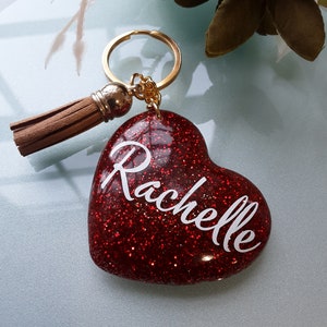 May include: Red glitter heart-shaped keyring with a gold key ring and a brown tassel. The heart has the name "Rachelle" written in white.