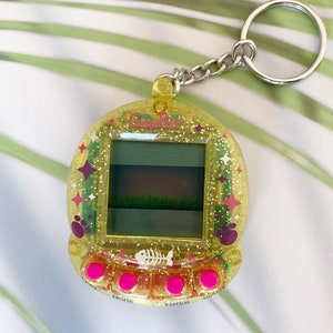 K&ouml;nnte beinhalten: Ein gelber digitaler Haustier-Schl&uuml;sselanh&auml;nger aus Kunststoff mit glitzernder Oberfl&auml;che. Der Bildschirm zeigt ein gr&uuml;nes Feld und einen wei&szlig;en Knochen. Der Schl&uuml;sselanh&auml;nger hat rosa Kn&ouml;pfe mit den Beschriftungen "Modus", "Eingabe" und "F&uuml;ttern".