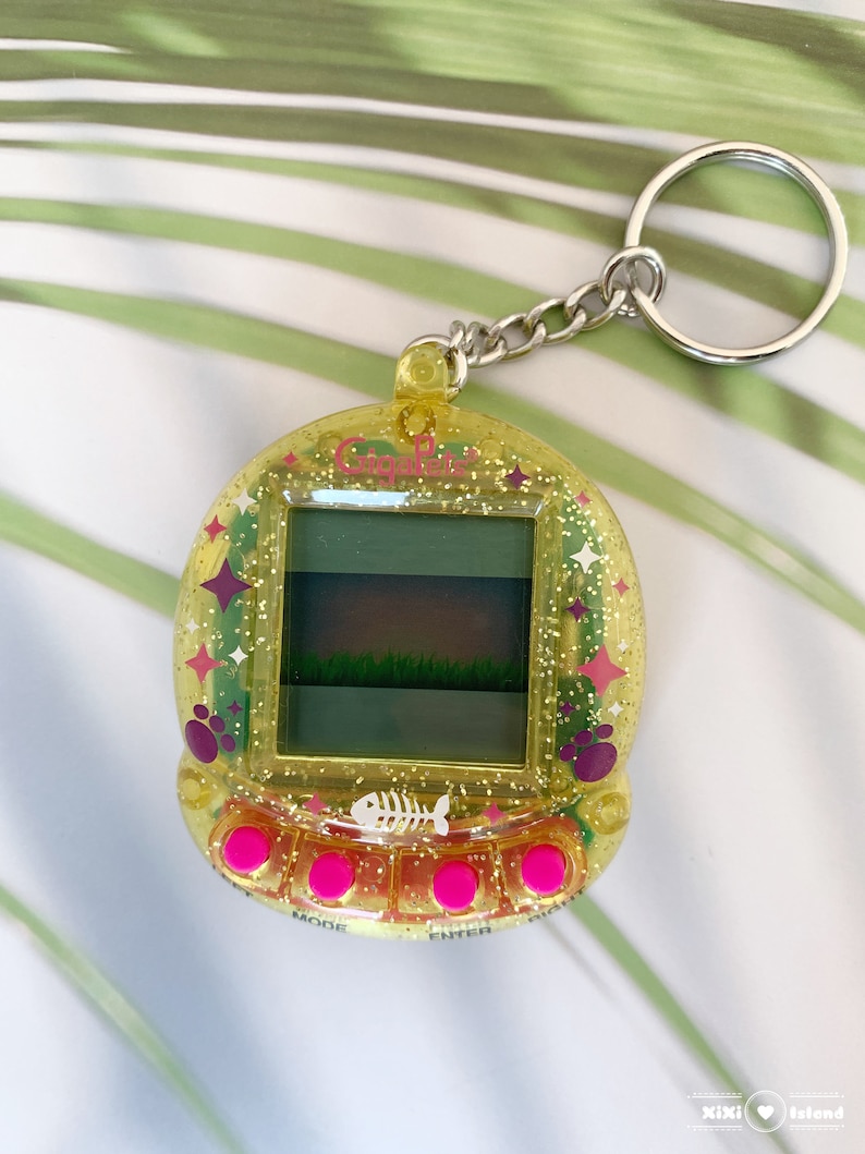 K&ouml;nnte beinhalten: Ein gelber digitaler Haustier-Schl&uuml;sselanh&auml;nger aus Kunststoff mit glitzernder Oberfl&auml;che. Der Bildschirm zeigt ein gr&uuml;nes Feld und einen wei&szlig;en Knochen. Der Schl&uuml;sselanh&auml;nger hat rosa Kn&ouml;pfe mit den Beschriftungen "Modus", "Eingabe" und "F&uuml;ttern".