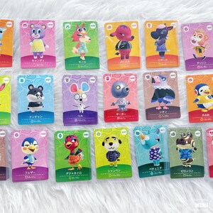 May include: A collection of Animal Crossing character trading cards. Each card features a different animal character with Japanese text and a unique design. The cards are arranged on a white, textured surface.
