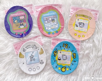 May include: Five colorful plastic plates featuring different Tamagotchi designs. The plates are round and have a white rim. The designs include a pink and blue Tamagotchi with a cat, a pink and green Tamagotchi with a bathtub, a yellow and pink Tamagotchi with a bunny, a white Tamagotchi with a snowflake, and a blue Tamagotchi with a clock.