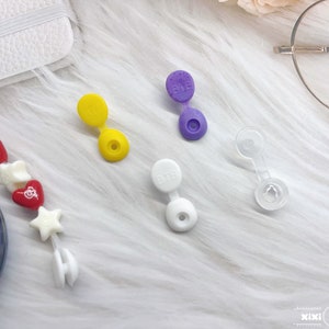 May include: Four plastic bottle stoppers in different colors: yellow, purple, white, and clear. The stoppers have a small loop on top and a flat bottom. The purple and white stoppers have the word "BTE" printed on them.