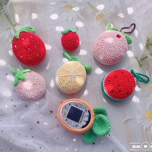Op de afbeelding: Een verzameling gehaakte sleutelhangers in de vorm van fruit, waaronder een aardbei, een watermeloen, een citroen, een perzik en een kleine rode aardbei. De sleutelhangers worden getoond op een wit oppervlak met een wit en zilveren gaasstof.