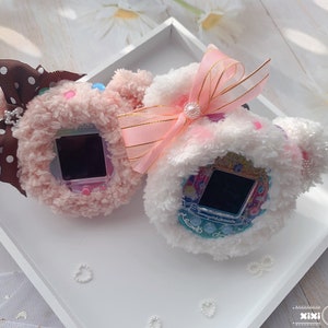 May include: Two plush toy cases for a Tamagotchi, one pink with brown polka dot bow and one white with pink bow. Both cases have a Tamagotchi inside.
