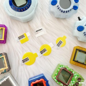 May include: A collection of colorful Digivice toys in various colors, including blue, white, purple, green, and yellow. The toys have rectangular screens and buttons, with some featuring protective screen covers labeled "FRONT".