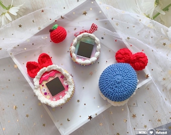 May include: Three crocheted Tamagotchi covers. One is pink and white with a red bow, one is blue and white, and one is white with red and white accents. The covers are on a white surface with a white lace overlay.