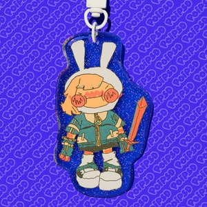 May include: A blue and white glitter keychain featuring a cartoon character with a bunny ear hat holding a sword. The character has blonde hair, blue eyes, and is wearing a blue and green jacket.