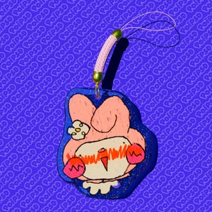 Puede incluir: Un amuleto de personaje azul y rosa con una flor en la cabeza. El amuleto está unido a una cuerda dorada y blanca.