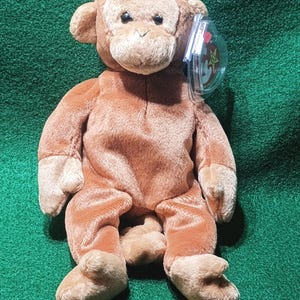 May include: A plush brown monkey with a soft, textured appearance. The stuffed animal has a light brown face, ears, hands, and feet. It has black eyes and a tag attached to its ear. The monkey is sitting on a green surface.