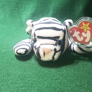 May include: White and black striped beanie baby tiger plush toy with blue eyes. The toy has a red and white Ty tag with the words "Beanie Original Baby" on it.