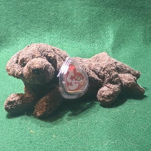 May include: A brown plush dog toy with a tag that reads "2000". The toy is lying down on a green surface. The dog has a soft, textured appearance.