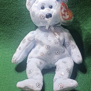May include: A light blue beanie baby bear with a silver snowflake pattern. The bear has a red and white Ty tag attached to its ear.